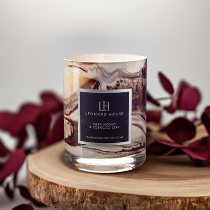 Dark Honey and Tobacco Scented Candle in brown swirl pattern glass on a wooden board with dark red flowers